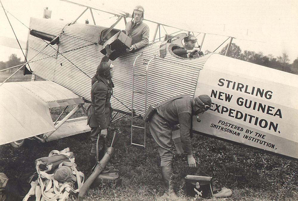 photo of men in old fashioned flying gear sitting in and loading a two-person prop plane