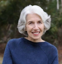 Headshot of a woman, smiling, with short, wavy white hair. She wears a blue sweater and there are trees in the background.