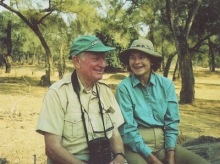 A older man and an older woman sit in the African savannah. The man has binoculars on his head and both are wearing hats and loose clothing.