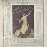 Cover of Columbia bicycle catalog, 1912, showing a woman kneeling painting the words Columbia while crowning a bicycle with a laurel wreath.